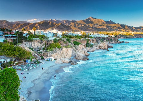 Aerial view of Nerja, Spain with Burriana Beach along the coastline, the Sierra Almijara mountains in the background, and historic buildings including the Church of El Salvador and Balcón de Europa