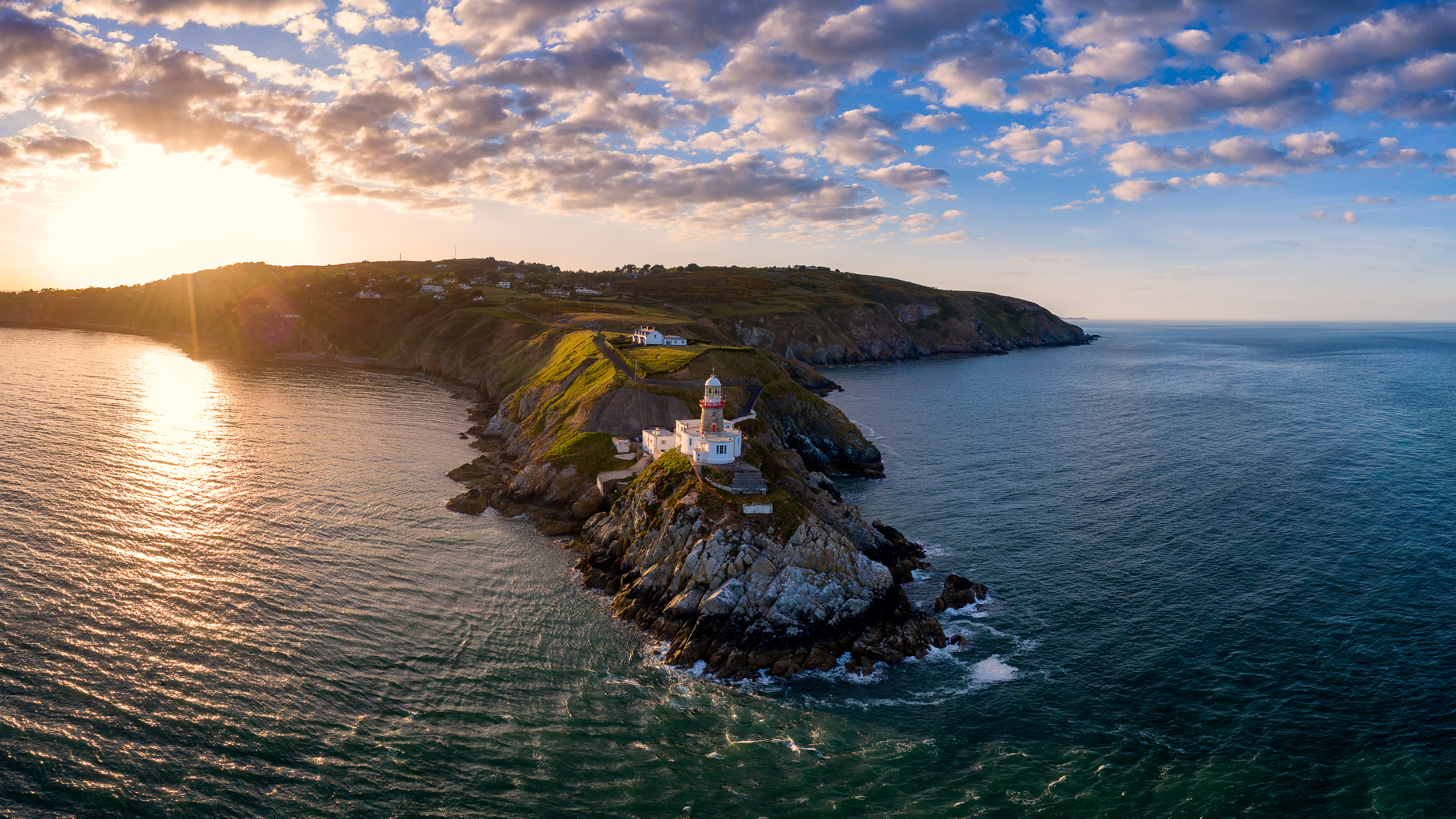 White lighthouse with a red band on rocky cliffs at Howth Head with green hills and houses above the coastline
