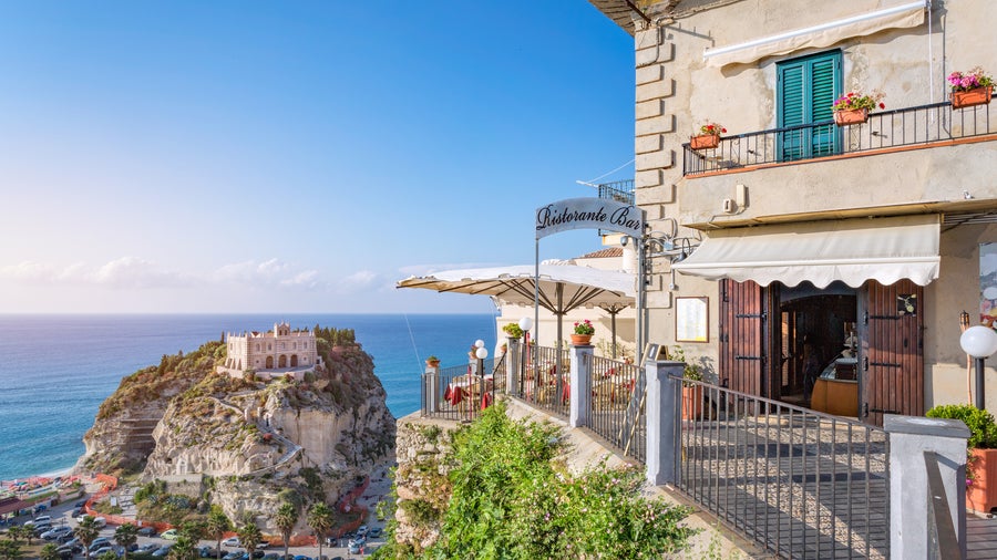 Traditional restaurant with classic balcony with Sanctuary of Santa Maria dell’Isola in the background at Tropea, Italy,
