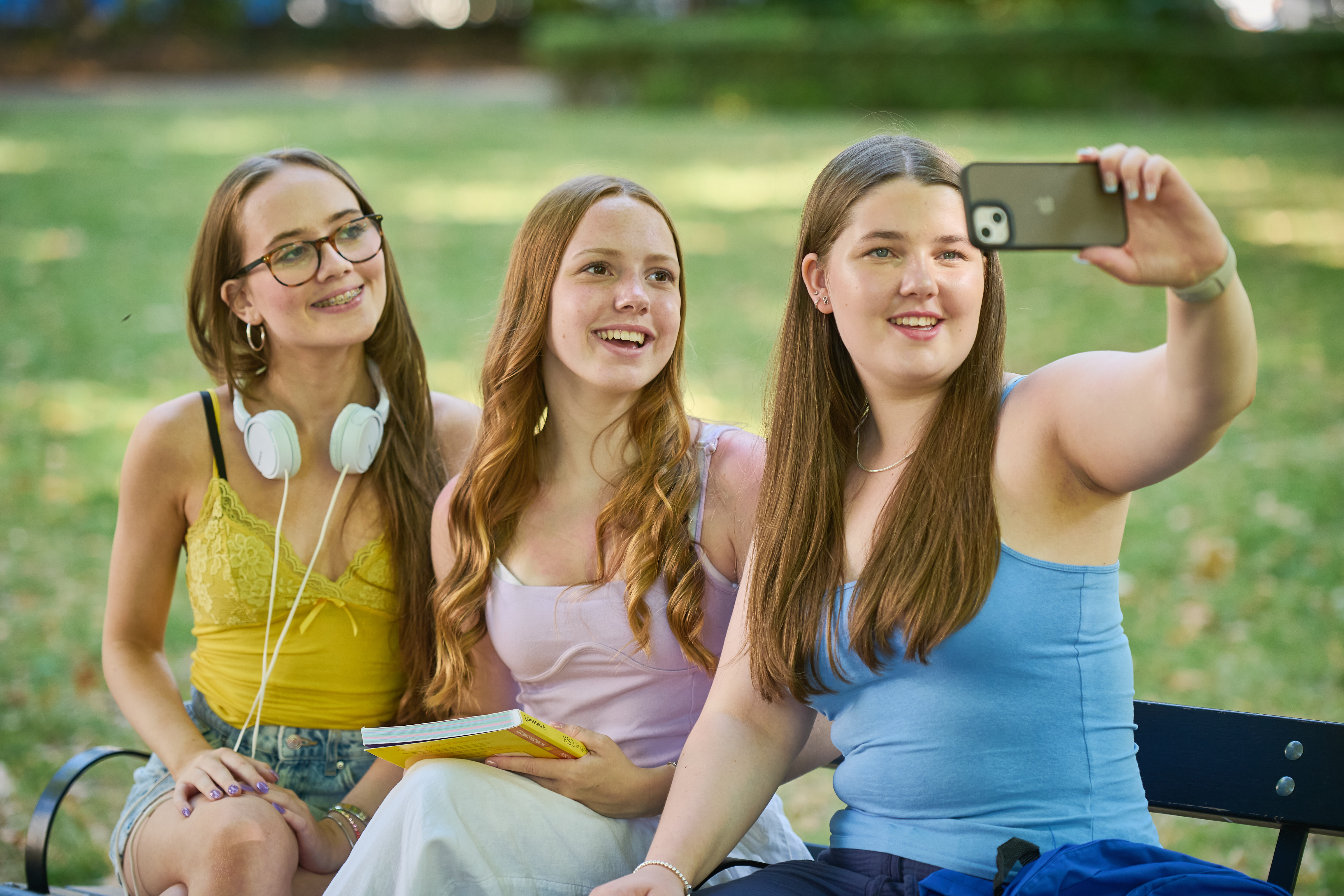 Three teen ESL girls sitting on bench taking selfies in London park, United Kingdom