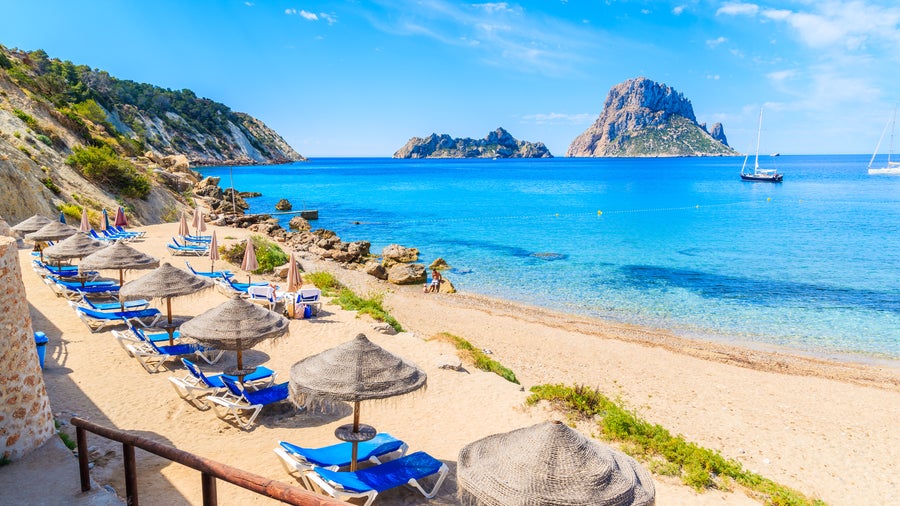 Beach at Ibiza, Spain with colorful umbrellas and beach chairs, crystal clear sea, and boats in the water