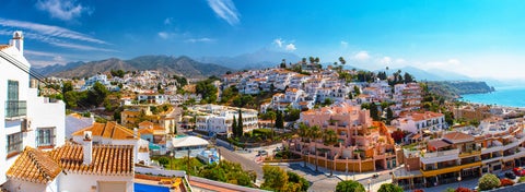 Aerial view of Nerja, Spain with Burriana Beach along the coast, Sierra Almijara mountains in the background, and historic sites like the Balcón de Europa and Iglesia El Salvador