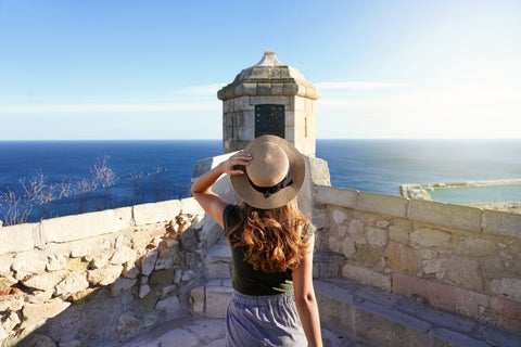 Tourist enjoying the seaside view from Santa Bárbara Castle in Alicante, Spain, overlooking El Postiguet Beach, the Mediterranean Sea, and the cityscape below