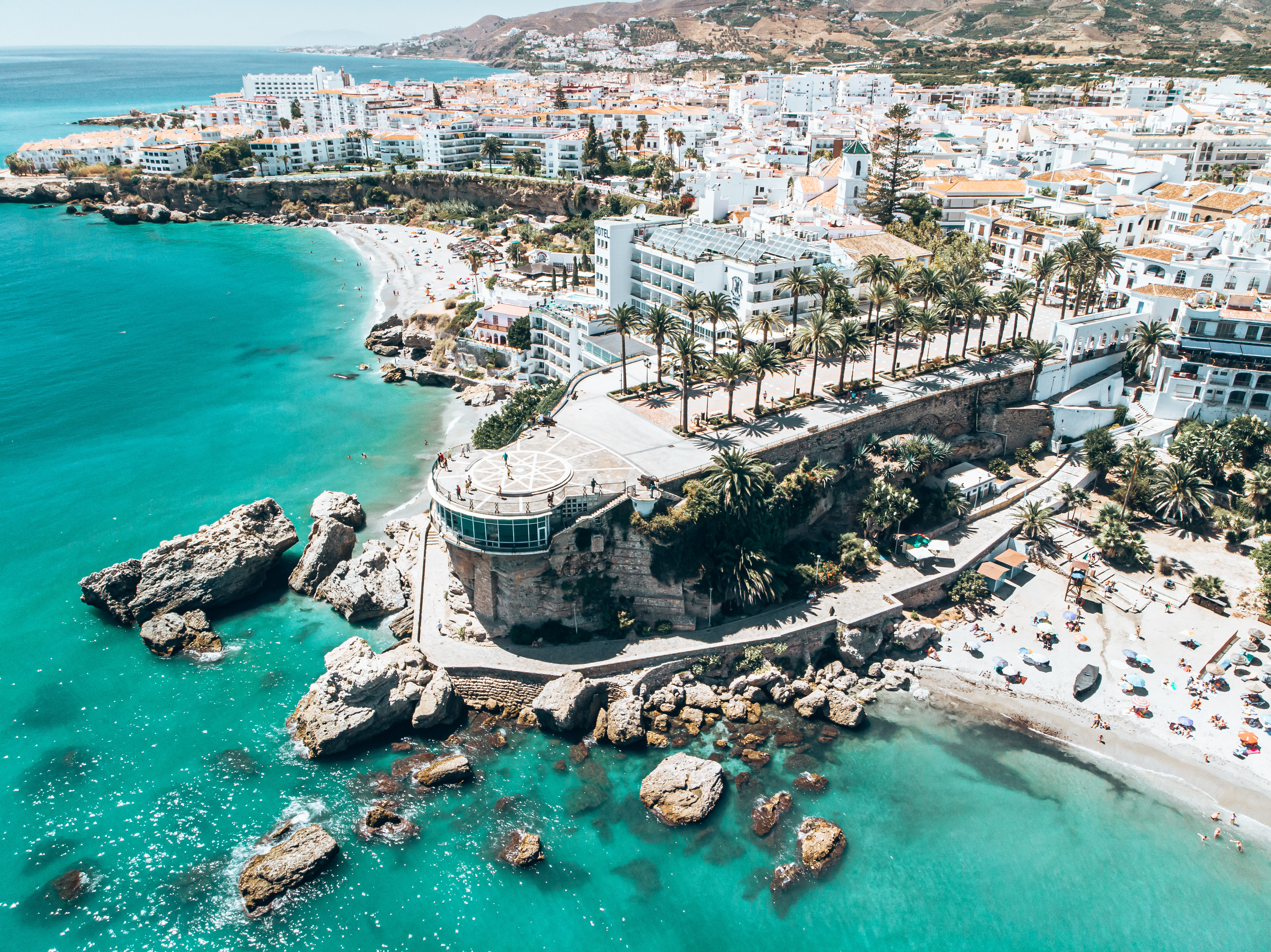 Aerial view of Nerja, Spain with Burriana Beach along the coastline, the Sierra Almijara mountains in the background, and historic buildings including the Church of El Salvador and Balcón de Europa
