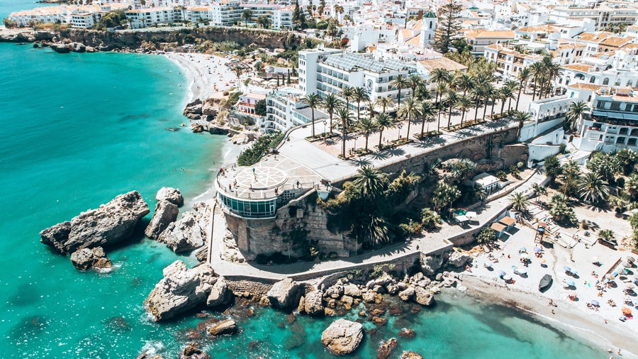 Aerial view of Nerja, Spain with Burriana Beach along the coastline, the Sierra Almijara mountains in the background, and historic buildings including the Church of El Salvador and Balcón de Europa