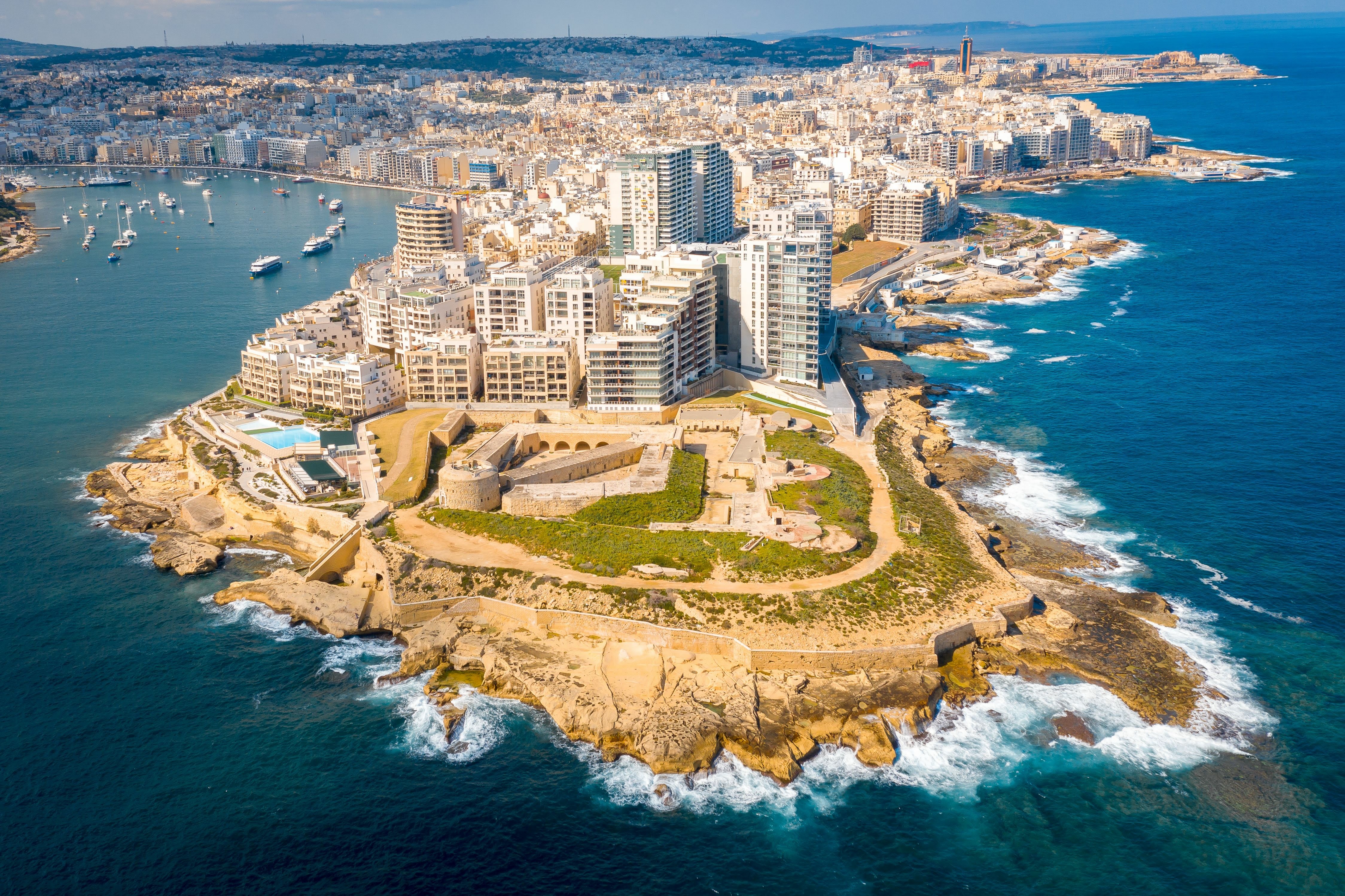 Aerial view of Sliema, Malta, surrounded by the Mediterranean Sea, showing boats in the harbor, modern buildings, and historic architecture