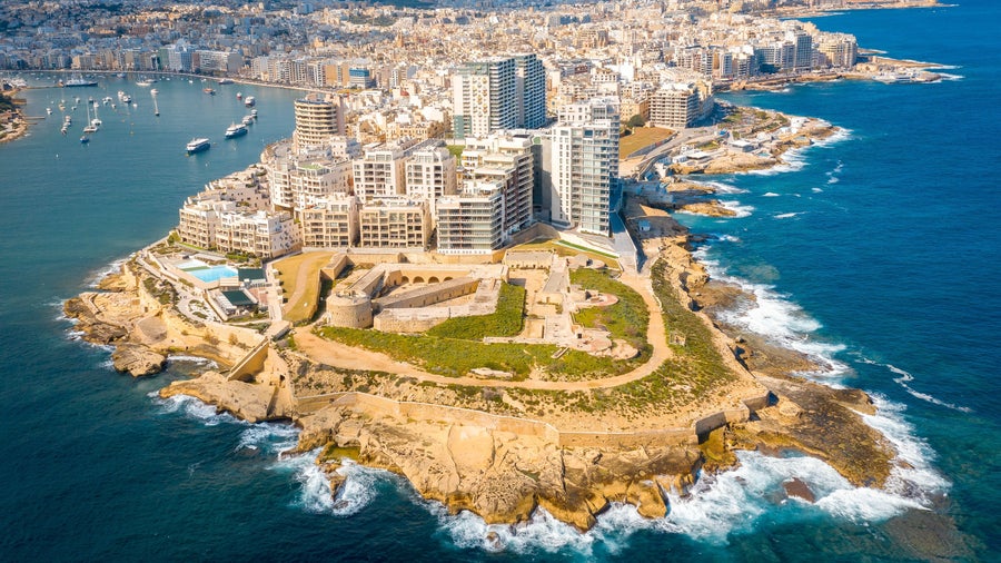 Aerial view of Sliema, Malta, surrounded by the Mediterranean Sea, showing boats in the harbor, modern buildings, and historic architecture