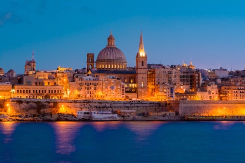 Historic skyline of Valletta, Malta, featuring golden limestone buildings, grand fortifications, domed churches, and the deep blue Mediterranean Sea at night