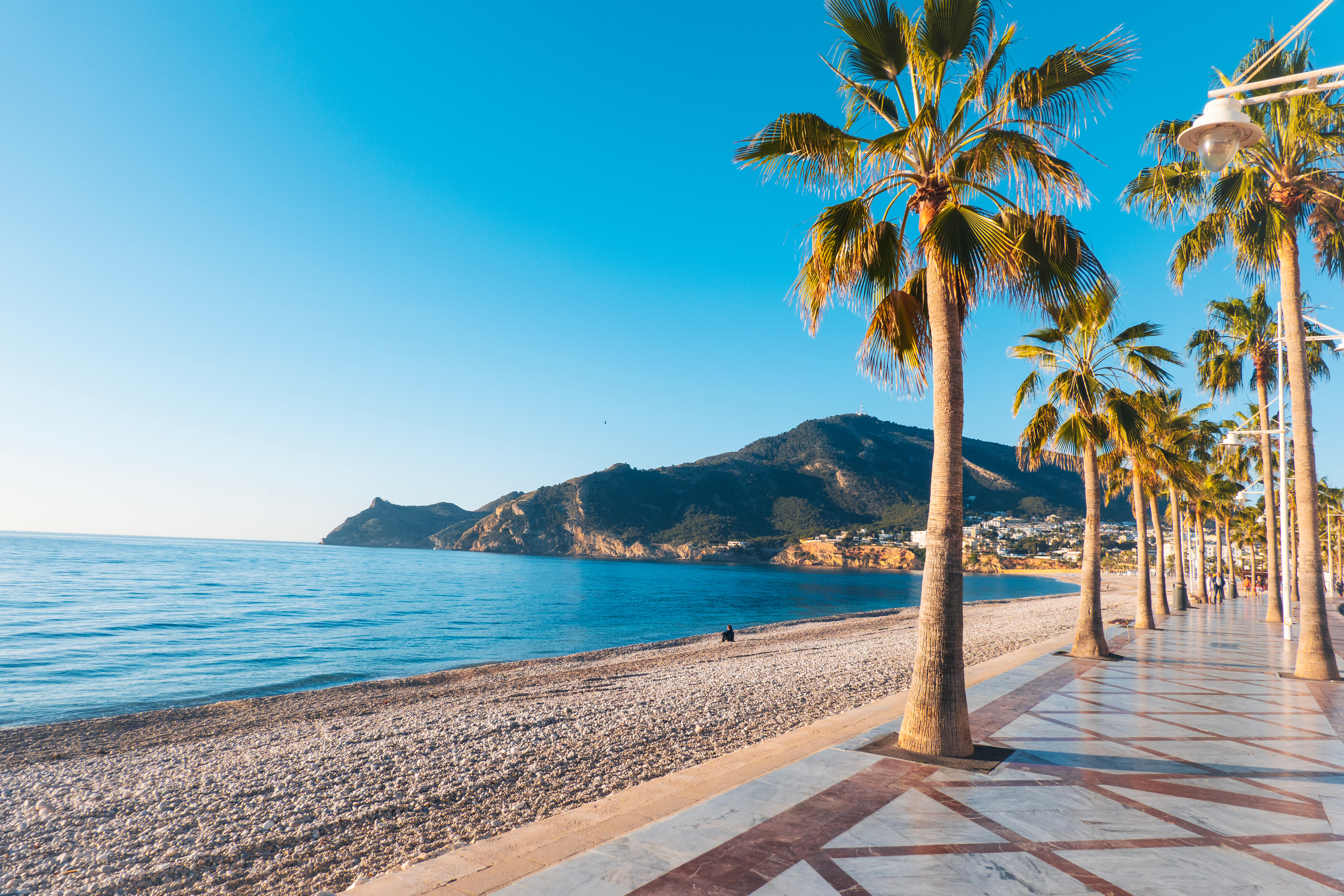 palm-lined promenade with a tiled sidewalk in Alicante, Spain, running alongside El Postiguet Beach with Mount Benacantil and Santa Bárbara Castle in the background