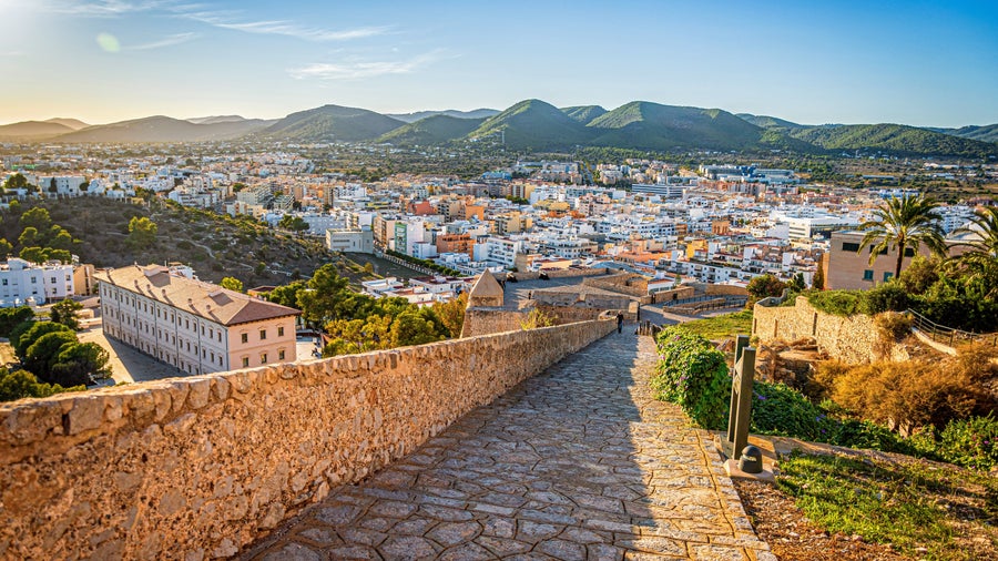 Aerial view of Ibiza, Spain with a rustic path descending towards the town, and mountains in the background