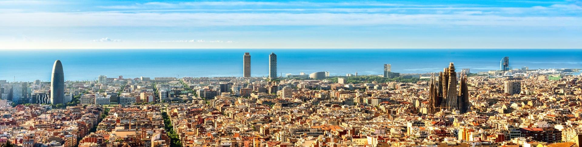 Aerial view of Barcelona, Spain, featuring the cityscape with buildings and streets stretching toward the Mediterranean Sea in the background