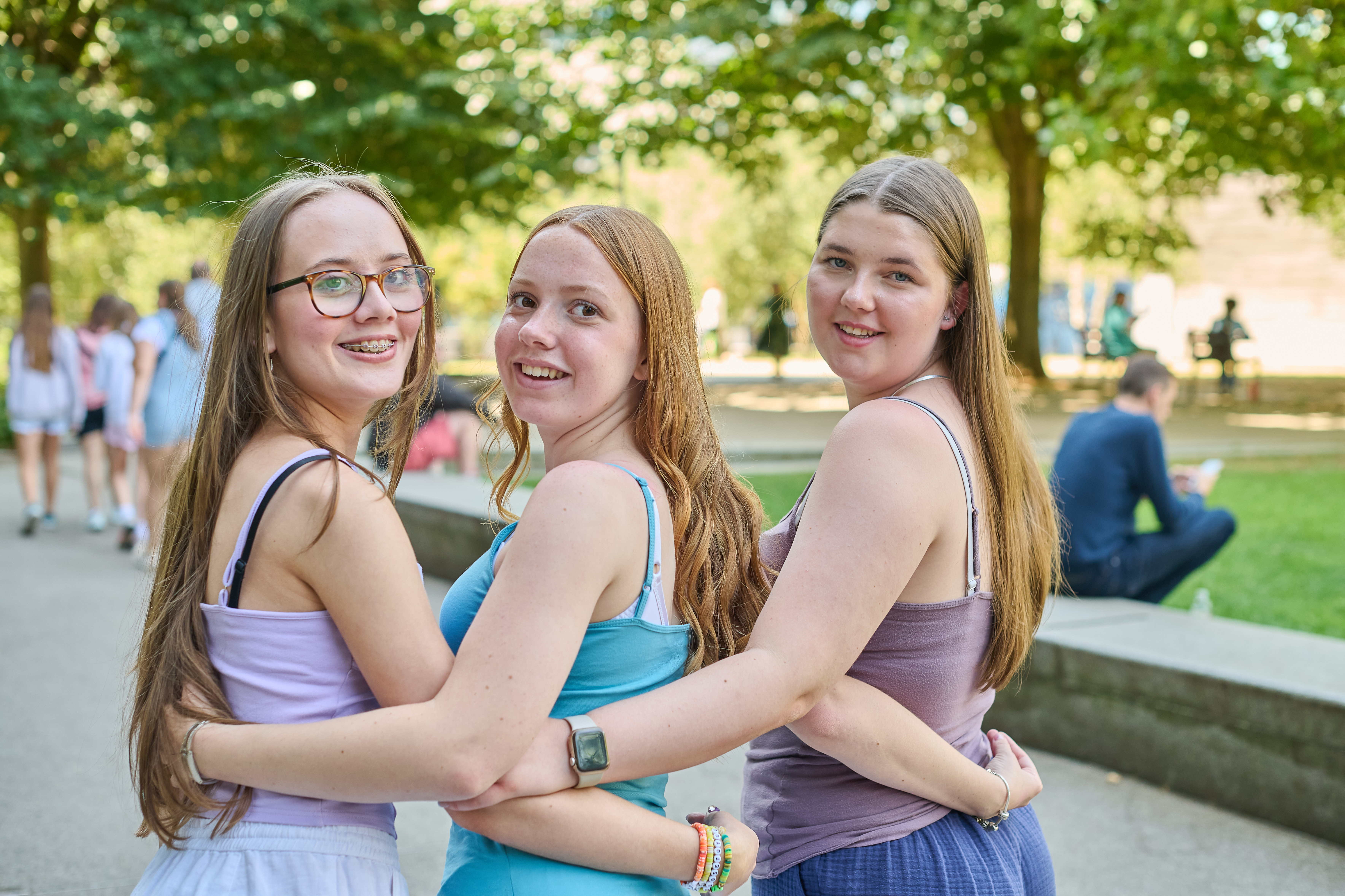 Three ESL teen girls smiling and holding each other in a bright green London park
