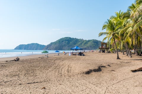 White-sand beach with beach umbrella, palm trees and Mountains in the back at Playa Jacó, Costa Rica