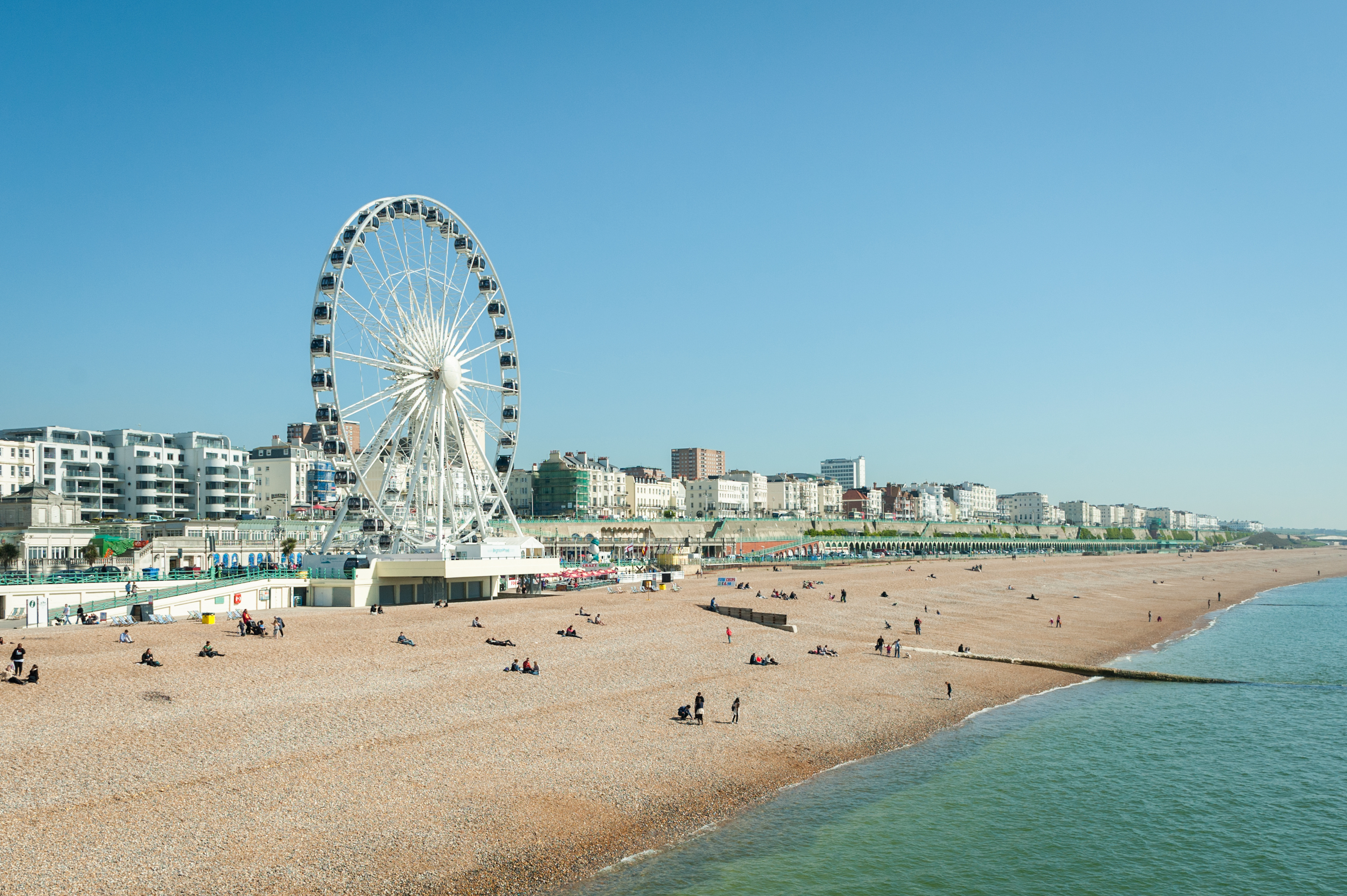 Brighton Beach, UK, people enjoying sunny day by the seafront with Ferris wheel, blue sky, and beach activities in the background