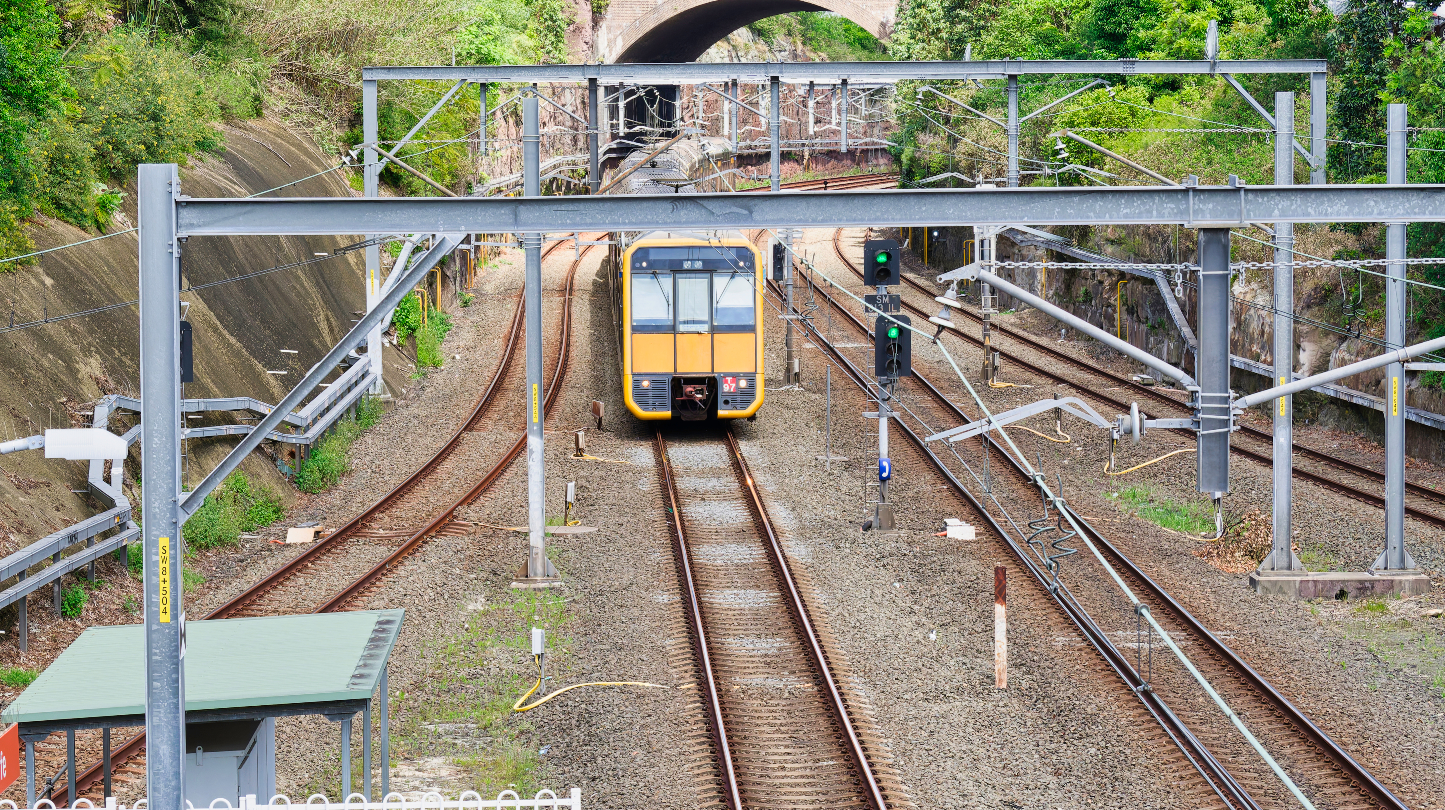 Yellow commuter train traveling on electrified railway tracks, with green signal lights and metal overhead structures, surrounded by vegetation and a stone tunnel in the background