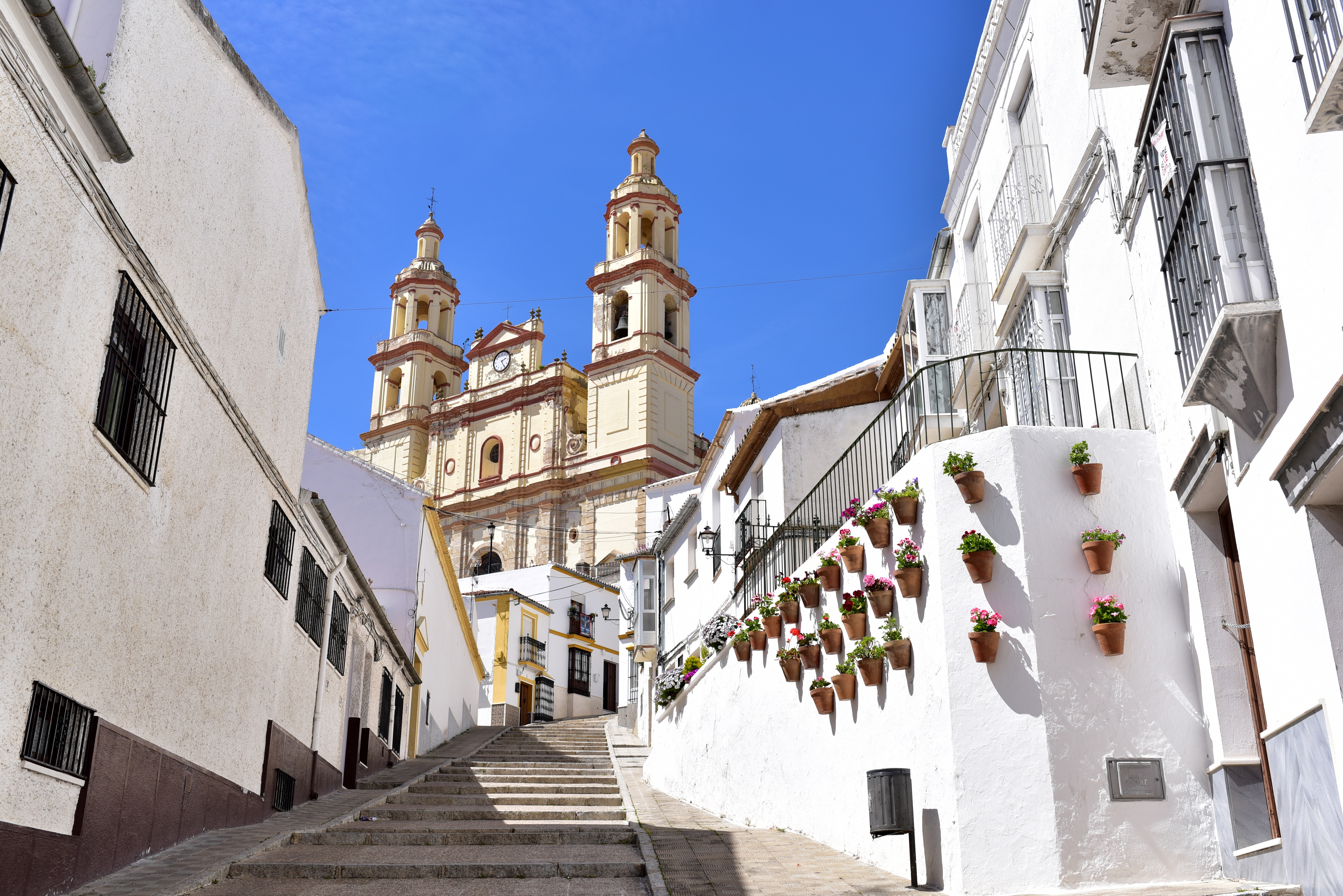 View of Cádiz Cathedral with its golden dome and cross, seen from a narrow street lined with traditional white buildings and palm trees