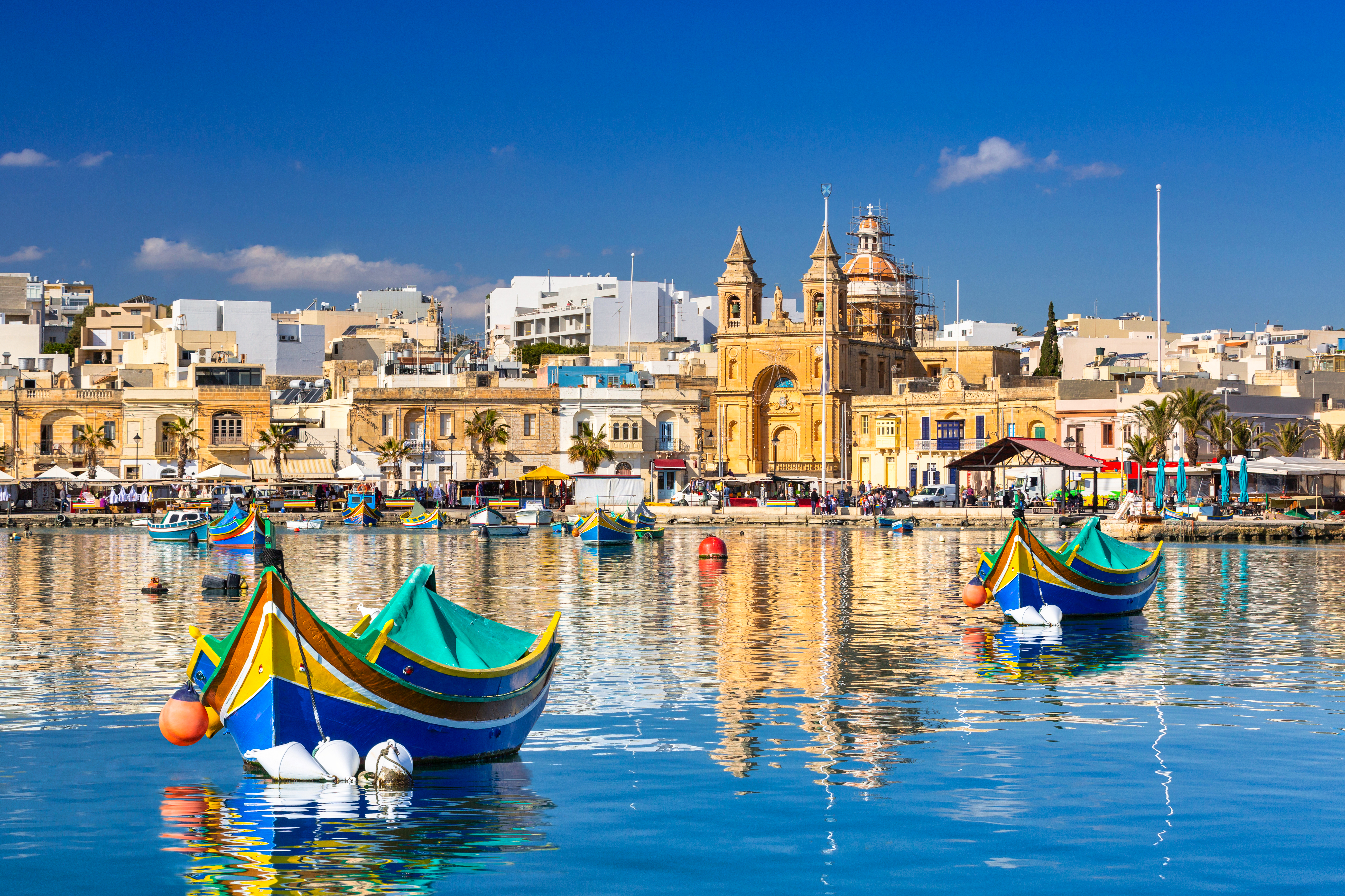 Colorful traditional Luzzu boats docked at Marsaxlokk Harbor in Malta, with historic stone buildings and modern apartments in the background
