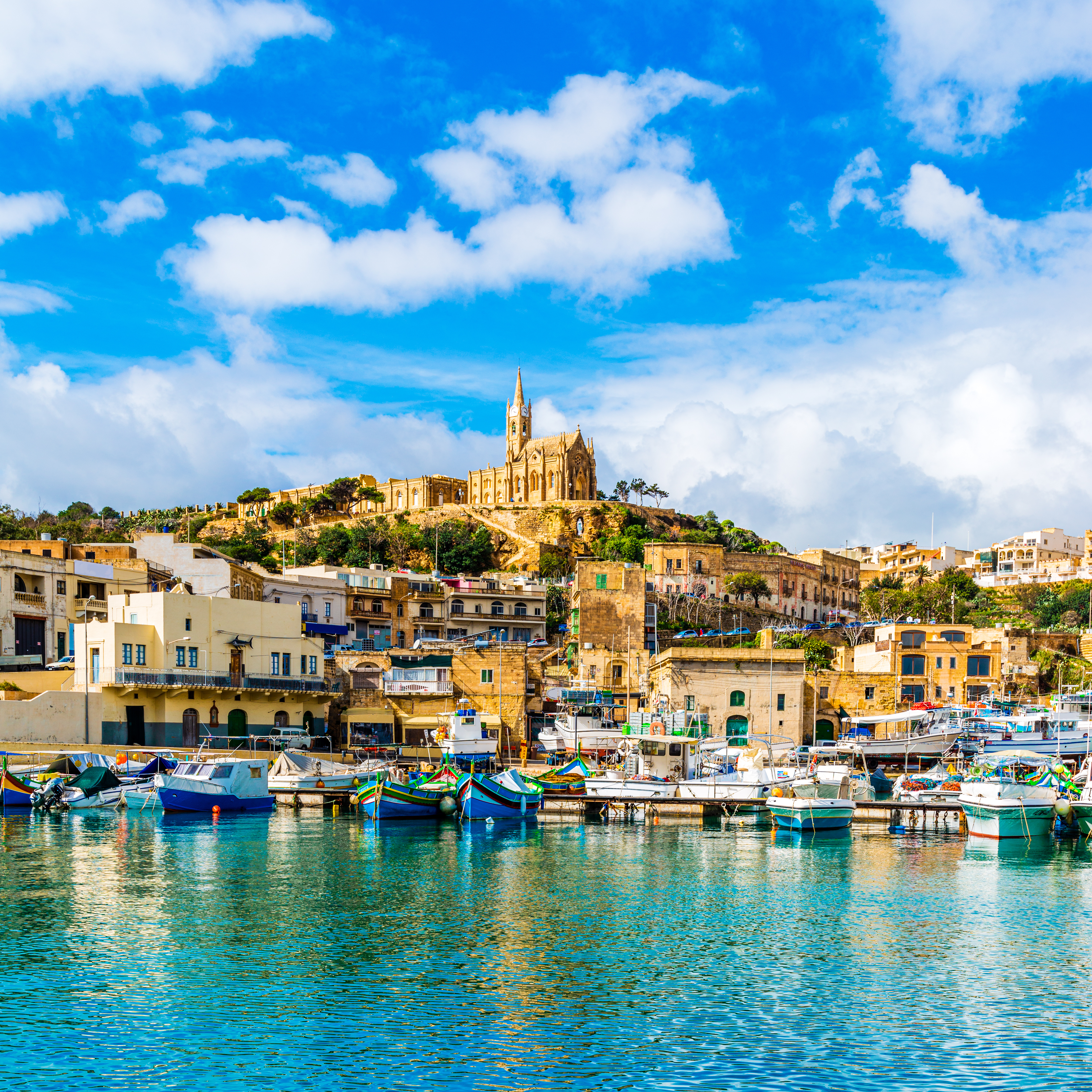 Boats anchored in the sea at the port of Mgarr, Gozo, Malta, with the historic Church of Our Lady of Lourdes overlooking from the hill in the background