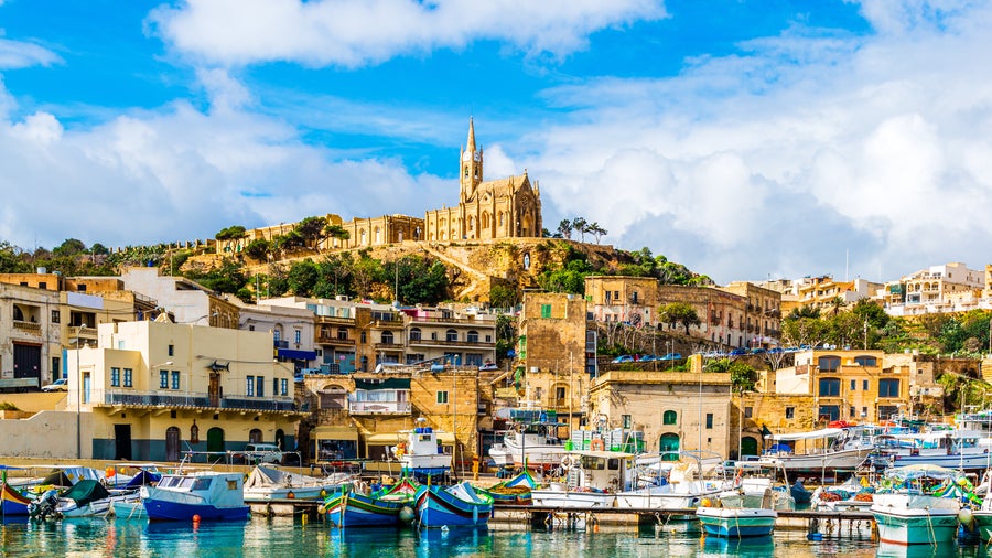 Boats anchored in the sea at the port of Mgarr, Gozo, Malta, with the historic Church of Our Lady of Lourdes overlooking from the hill in the background