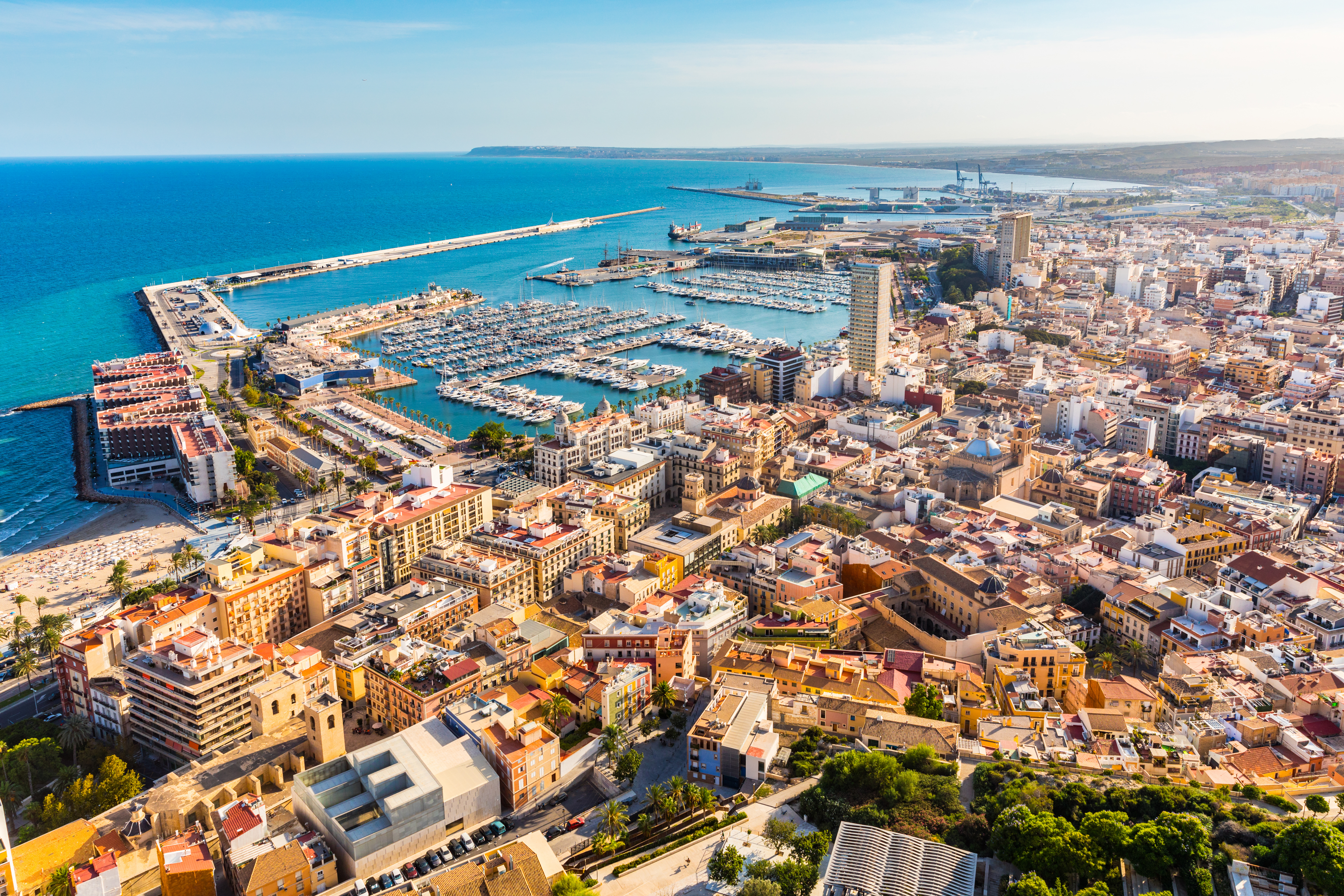 Panoramic aerial view of Alicante, Spain, showing El Postiguet Beach, Santa Bárbara Castle on Mount Benacantil, colorful cityscape, marina with boats, and Mediterranean coastline