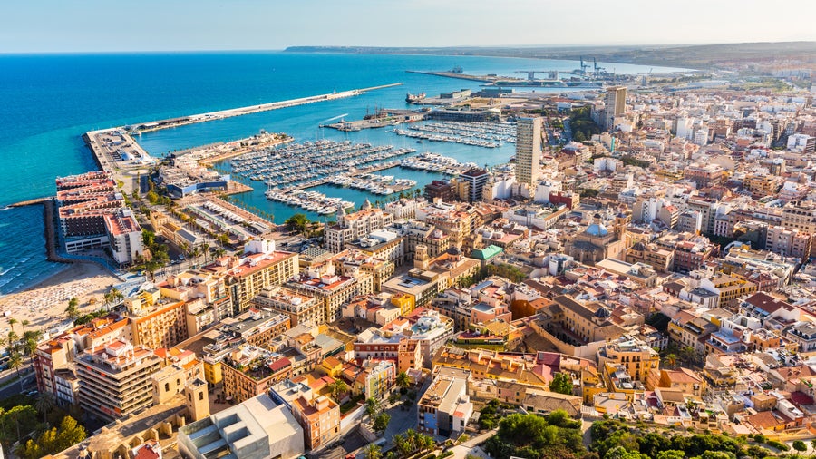Panoramic aerial view of Alicante, Spain, showing El Postiguet Beach, Santa Bárbara Castle on Mount Benacantil, colorful cityscape, marina with boats, and Mediterranean coastline