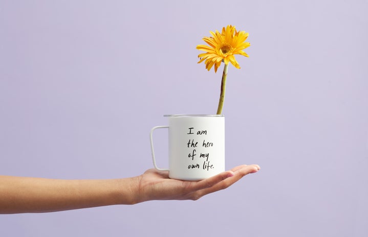 Hand holding white mug with “I am the hero of my own life” written on it and yellow flower sitting in it.
