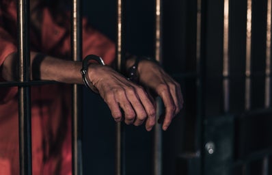 The image shows a person in an orange jump suit behind jail cell bars, with handcuffs on their wrist poking through the bars.