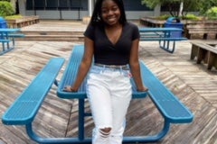 Young Black woman, Sheila Hodges, leaning against an aqua blue table and wearing a black top, white jeans with a belt, and sandals.