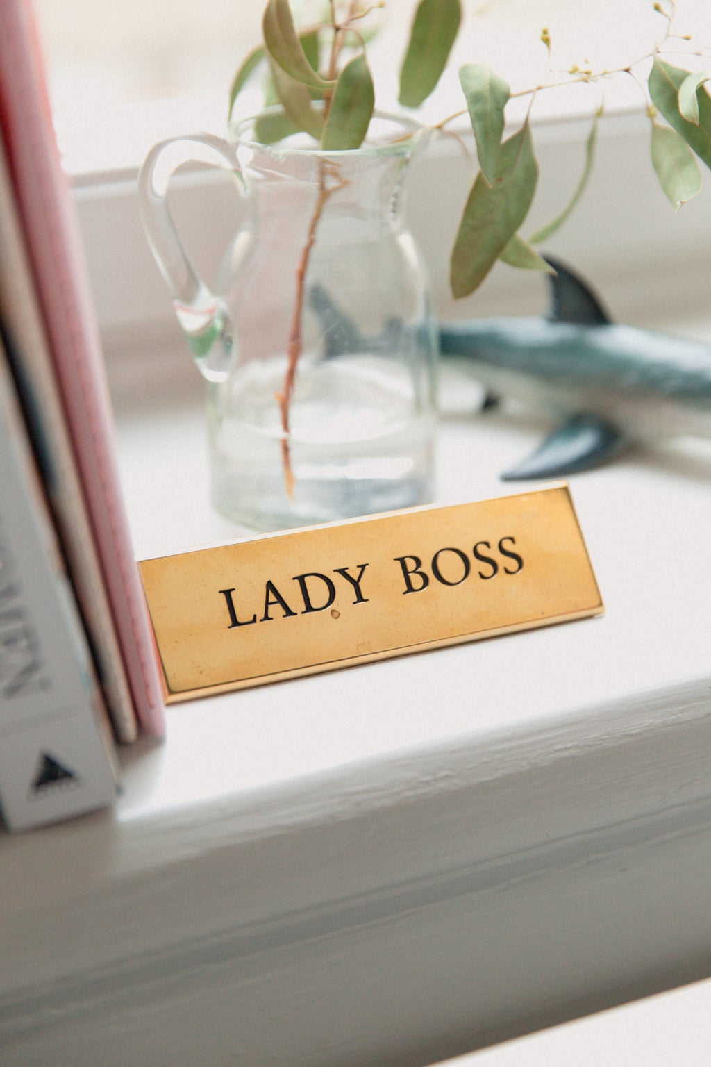 minimalist desk space with a pink folder and a plant. a gold plaque reads "lady boss"