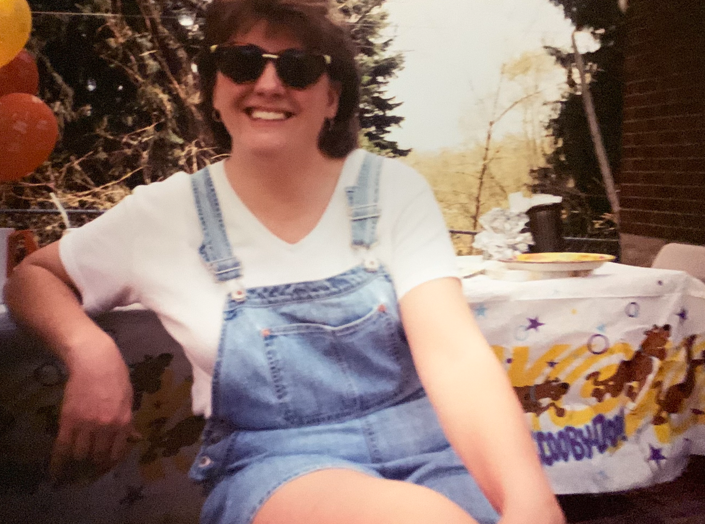 Woman smiling wearing overalls and large sun glasses.
