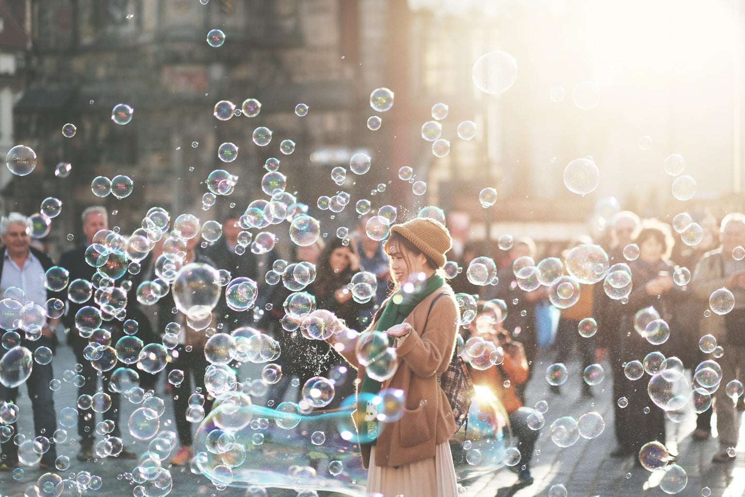 Woman outdoors in a beige coat and hat playing with bubbles, a crowd of folks in the background