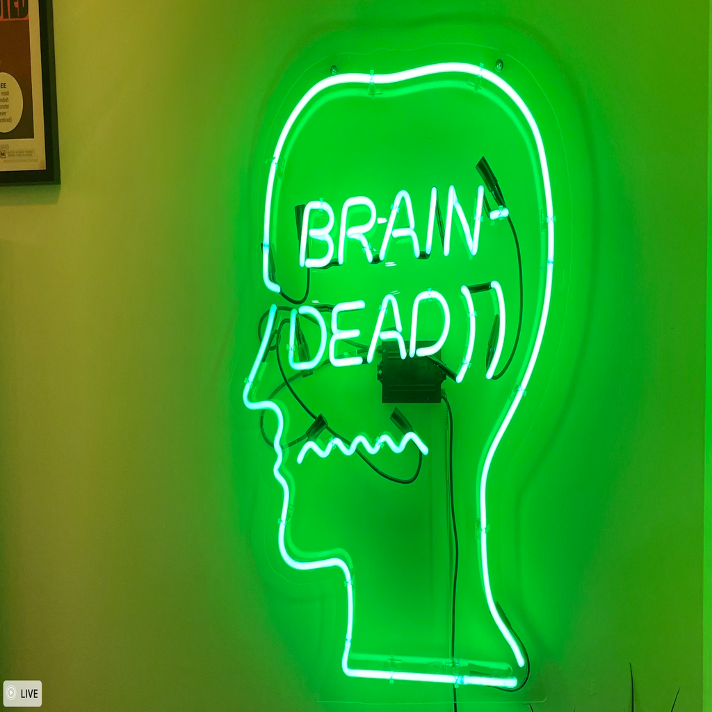 Neon green Brain Dead sign inside an outline of someone\'s side profile, hung on a wall.