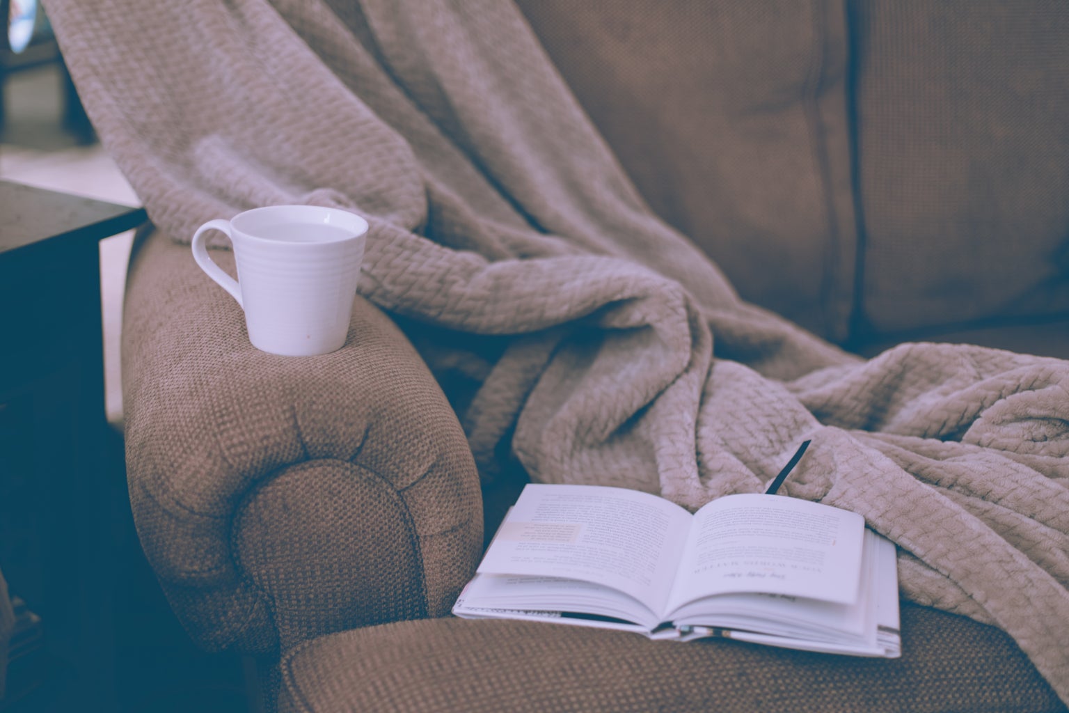 A book, a white ceramic mug and a blanket on a couch.