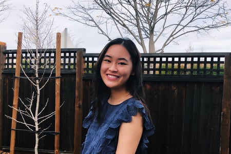 Girl in a blue top is smiling in front of a fence