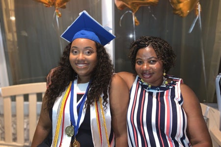 a mother and daughter, who is a graduate, sitting down on a bench in front of a window and smiling at the camera.