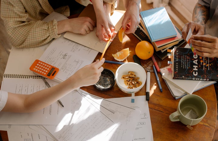 An image of a table top covered in snacks and paperwork. Four sets of hands work over it, sharing pencils and the food on the table in a comfortable manner.