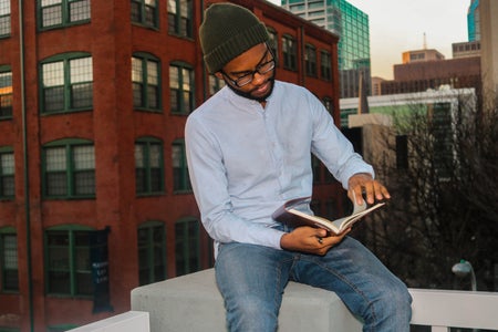 Black Poet Gabriel Angrand sitting, wearing glasses, in blue jeans, light-blue button-up, and green hat, holding a book in his hands.