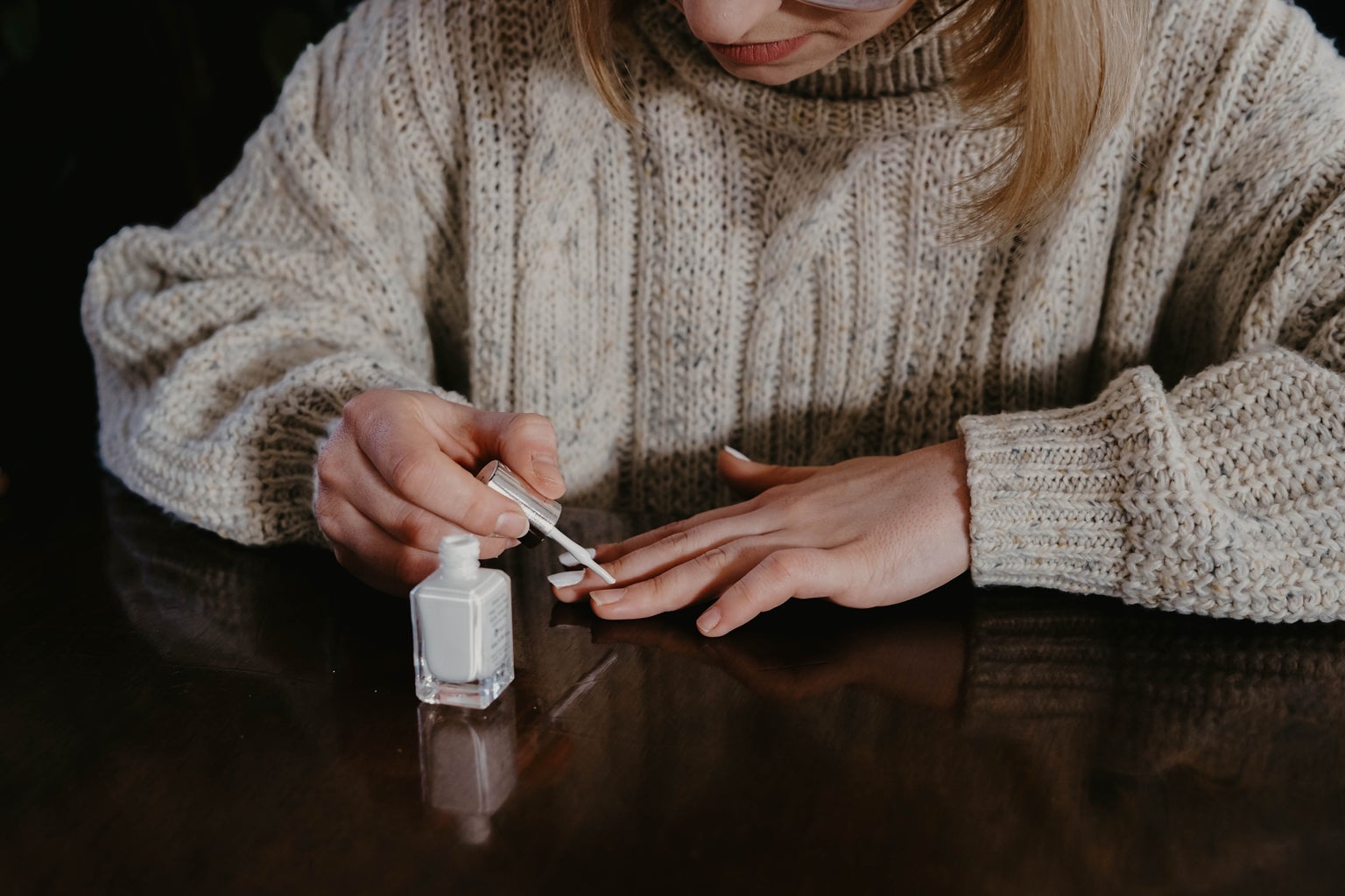 Woman in cream sweater painting her nails white on a table