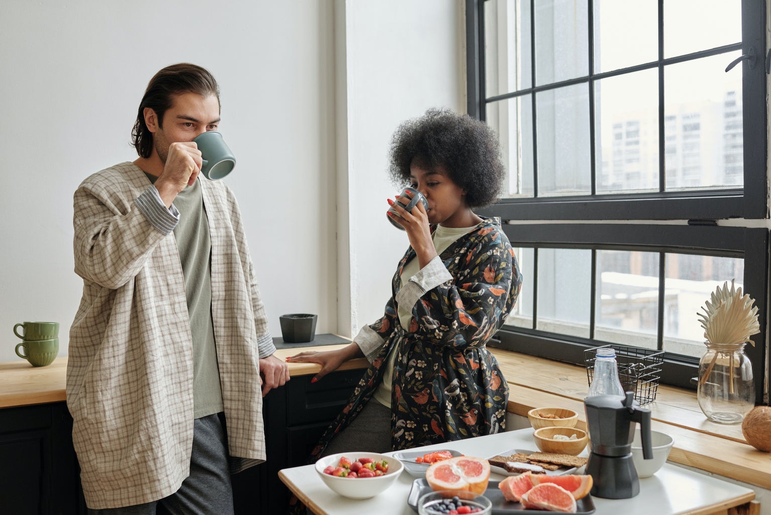 A couple standing in a brightly lit apartment drinking coffee. There is a table of fruit in front of them and they are in front of a window.