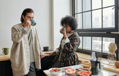 A couple standing in a brightly lit apartment drinking coffee. There is a table of fruit in front of them and they are in front of a window.