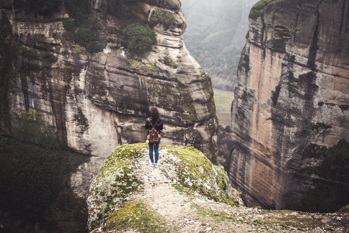 A woman with a red/brownish backpack is standing on a pathway on a cliff. She is facing the sides of two mountains.