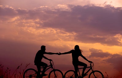 A woman and a man on bikes, reaching for each other with a sunset in the background