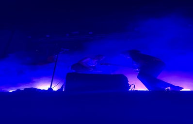 The image shows two people playing guitars on a stage with smoke and blue lights surrounding them.