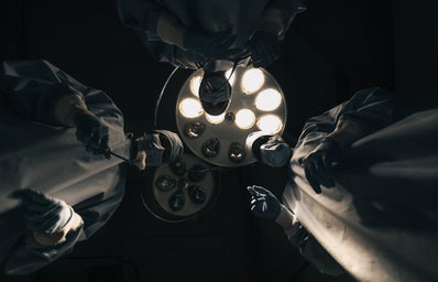 A point of view shot from an operating table staring up at three surgeons backlit by powerful operating lights.