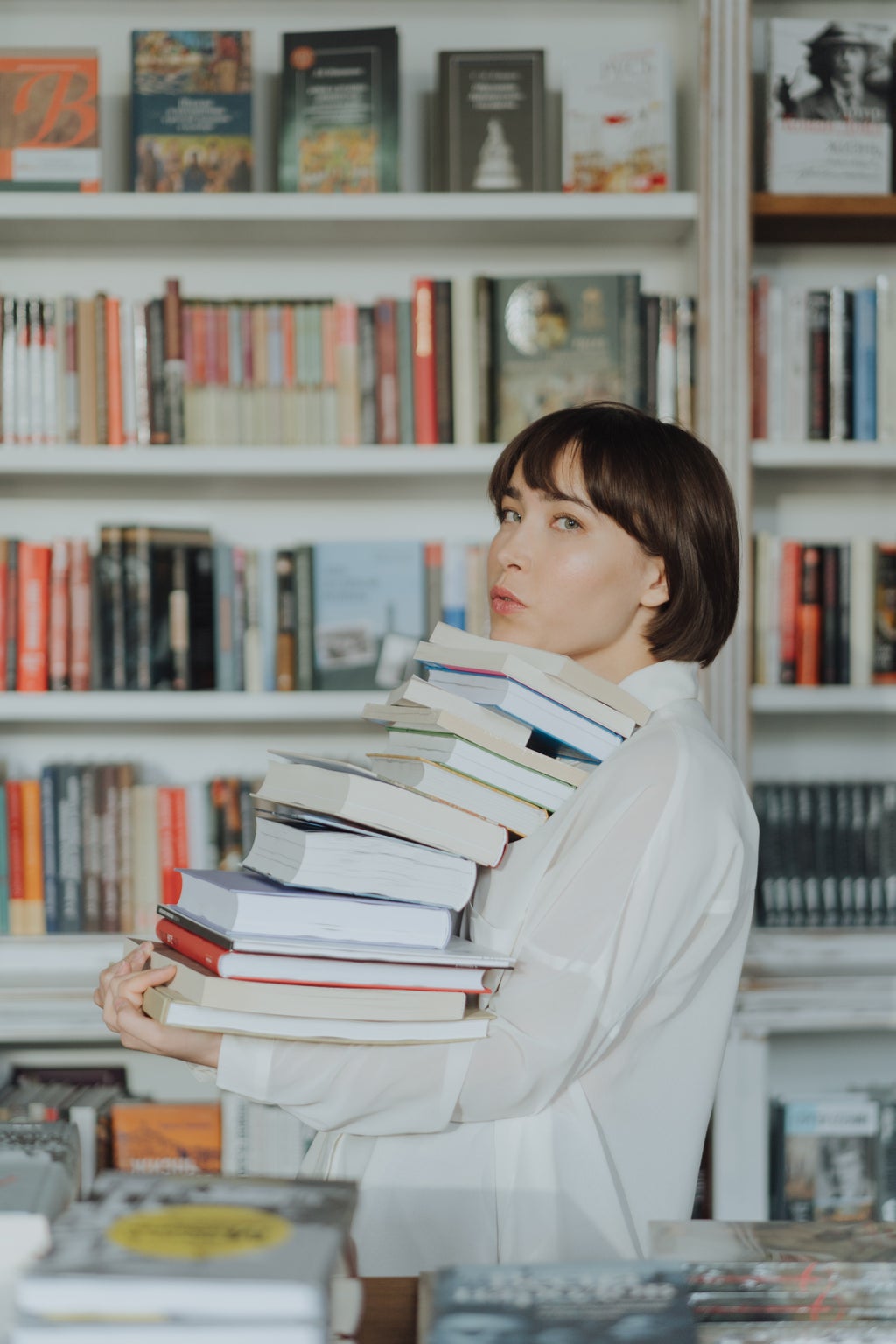 A woman in a white shirt carries a tall stack of books.