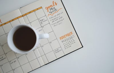 Coffee in a cup placed on top of a notebook. The notebook is lined with a calendar that has been filled out with events.