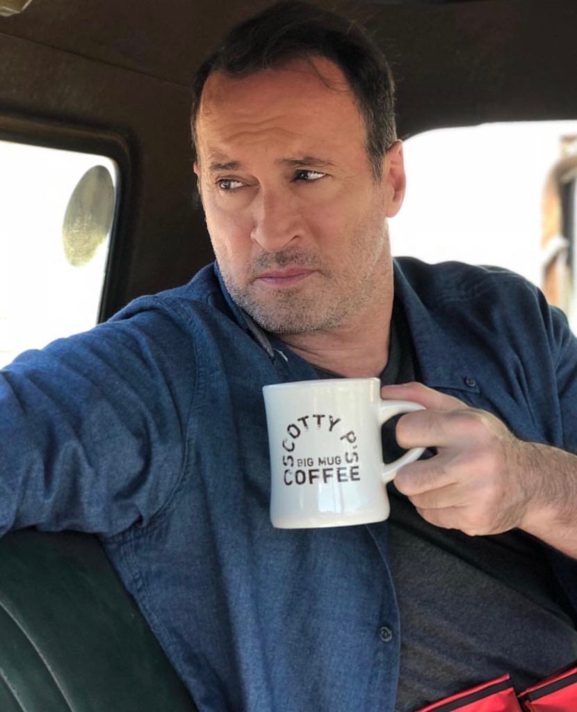 Image of Scott Patterson sitting in a car holding a cream-coloured coffee mug with the words \"Scotty P\'s Big Mug Coffee\" written on it