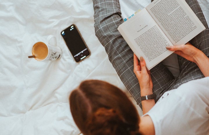 Woman sits in bed with her phone, a book and a mug.