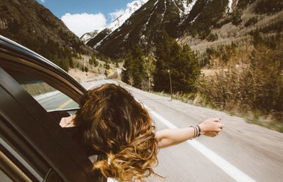 A girl with her head and hand out of a car window as it travels down a road with the backdrop of mountains