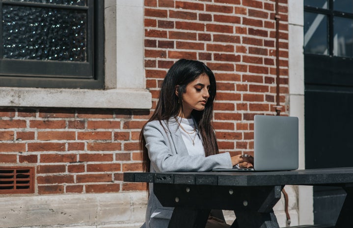 Woman sitting in front of a brick wall at a table with her laptop in front of her.